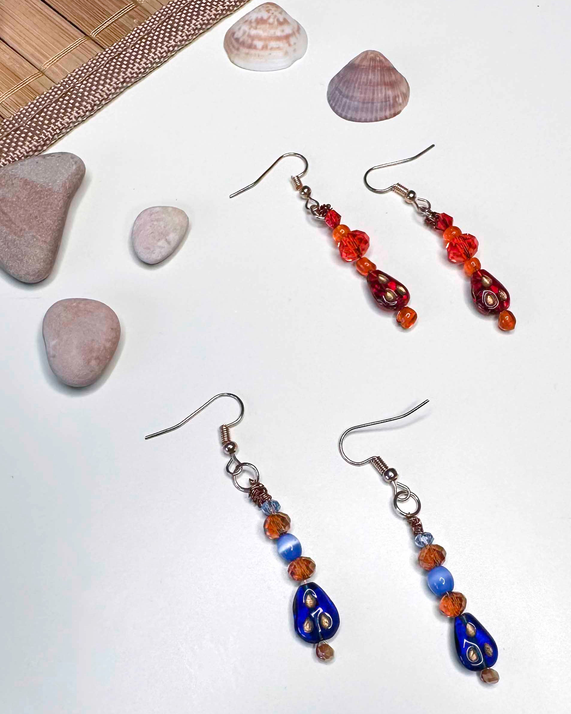 Two matching pairs of handmade dangle drop earrings, a red and orange beaded pair with red glass charms, and an orange and blue beaded pair with blue glass charms.