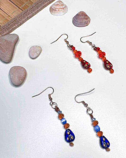 Two matching pairs of handmade dangle drop earrings, a red and orange beaded pair with red glass charms, and an orange and blue beaded pair with blue glass charms.