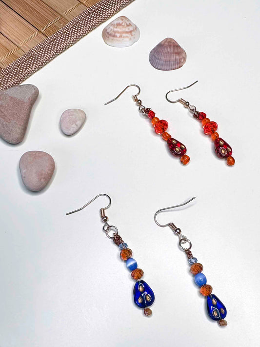 Two matching pairs of handmade dangle drop earrings, a red and orange beaded pair with red glass charms, and an orange and blue beaded pair with blue glass charms. 