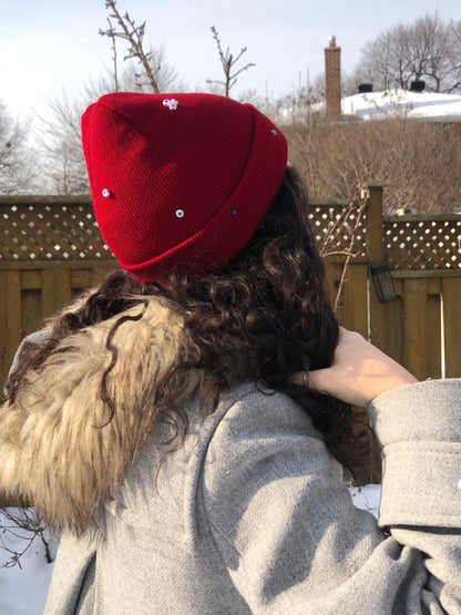 A red wool knit beanie decorated with both multicolor glass and circle heart beads, and an evil eye charm.