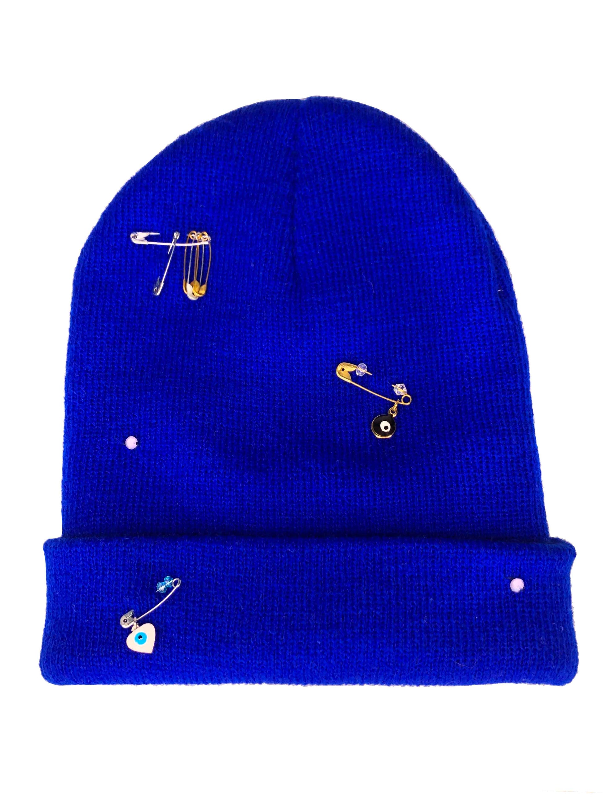 A blue wool knit beanie decorated with safety pins, evil eye charms and crystal beads.
