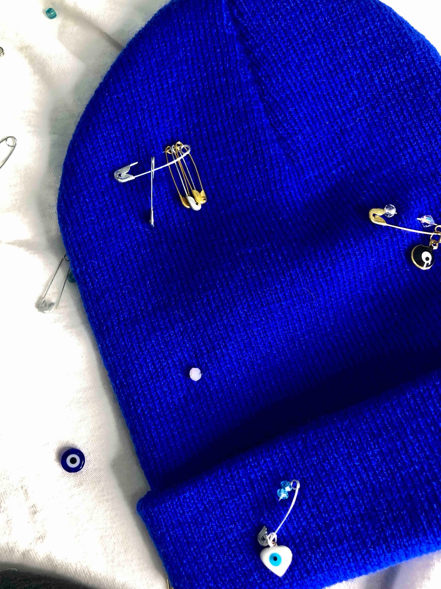 A blue wool knit beanie decorated with safety pins, evil eye charms and crystal beads.
