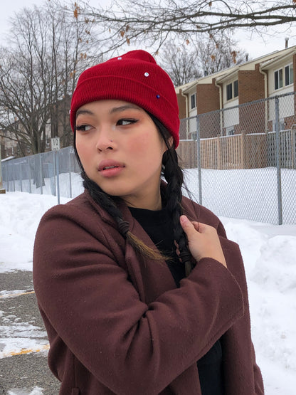 A red wool knit beanie decorated with both multicolor glass and circle heart beads, and an evil eye charm.